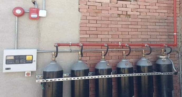 أسطوانات CO2 موصولة بمجمّع أحمر يصعد على الحائط، ولوحة التحكم إلى جانبها، وفوقها جرس إنذار وجهاز إنذار أحمر.