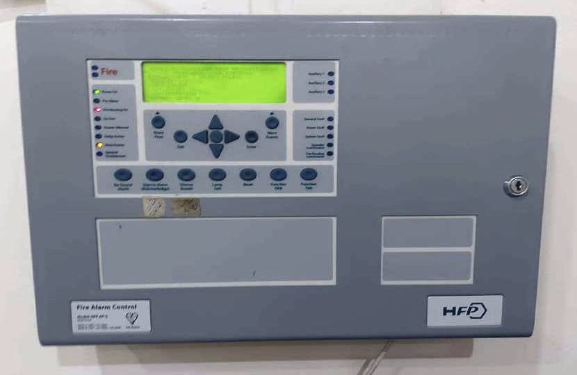 لوحة إنذار حريق رمادية بشاشة عرض نصية (LCD) وأزرار تحكم ومؤشرات حالة، مثبتة على الحائط.