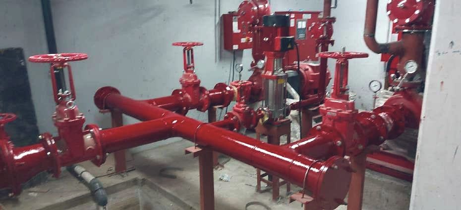 غرفة طلمبات مكتملة: مجمّع أحمر بمحابس بوابة (gate valves)، وطلمبة جوكي رأسية، ولوحات التحكم، ومقاييس الضغط.