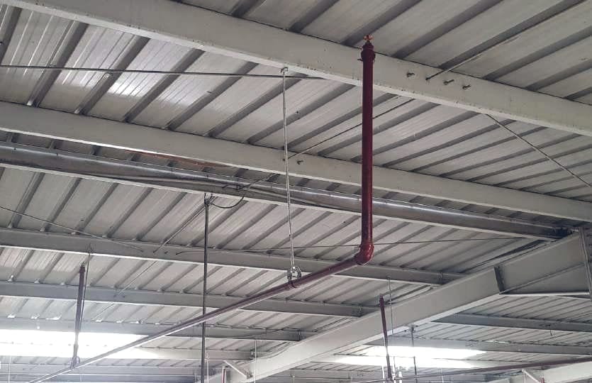 رشاش (sprinkler) قائم على وصلة حمراء من الخط الفرعي، تحت سقف معدني بجمالونات حديدية في مخزن، مع قضبان التعليق.